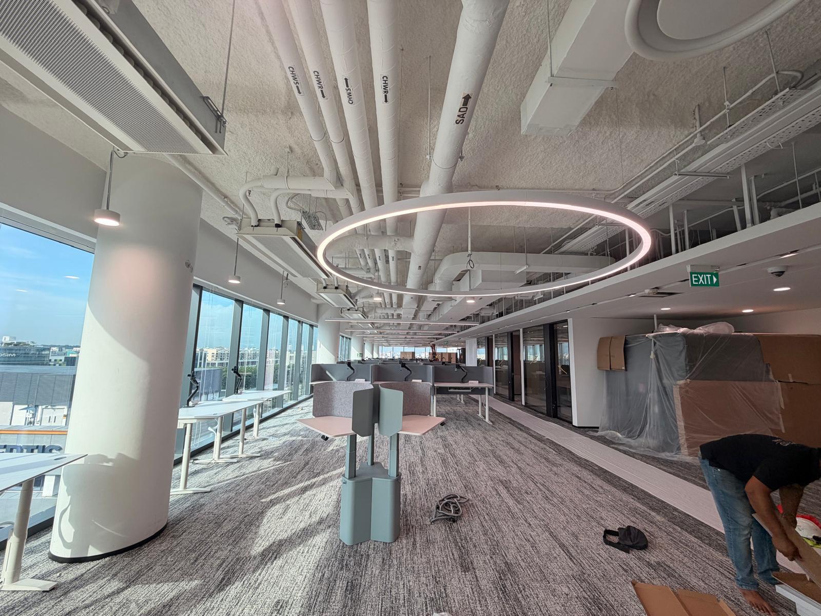 Circular suspended feature LED light installed above an open office workspace with exposed services, Singapore